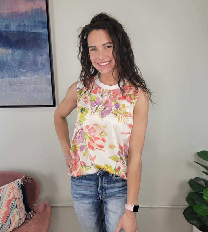 A woman with long brown hair, wearing the Staccato Carina Floral Blouse and blue jeans, smiles indoors near a pink chair, a framed painting, and a green plant.