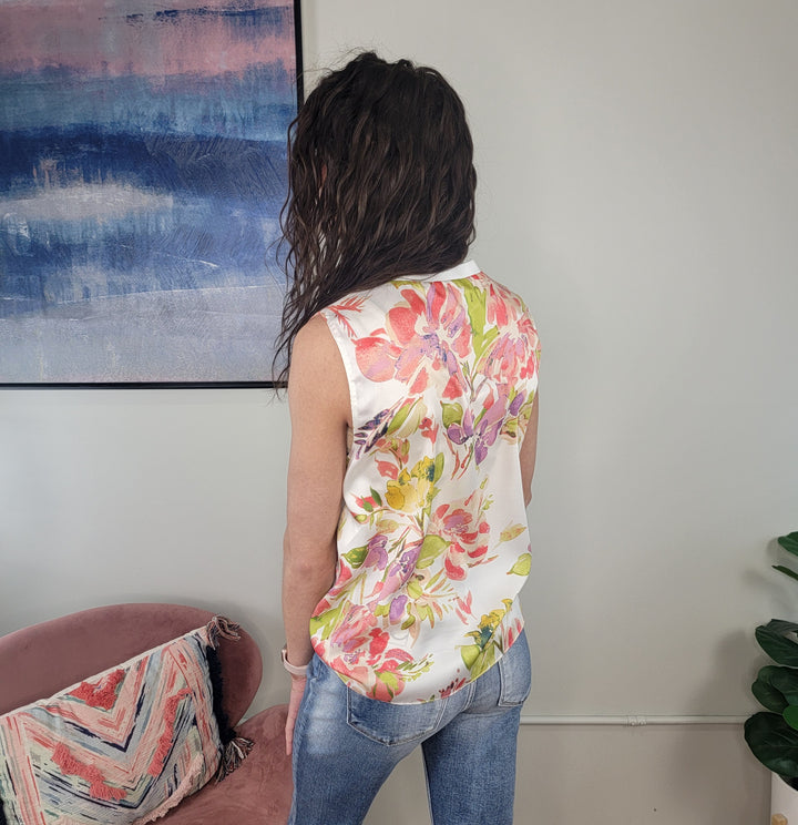 A person with wavy brown hair, wearing the Staccato Carina Floral Blouse and light blue jeans, stands with their back to the camera in a modern room featuring abstract art, a pink chair, and a green plant.