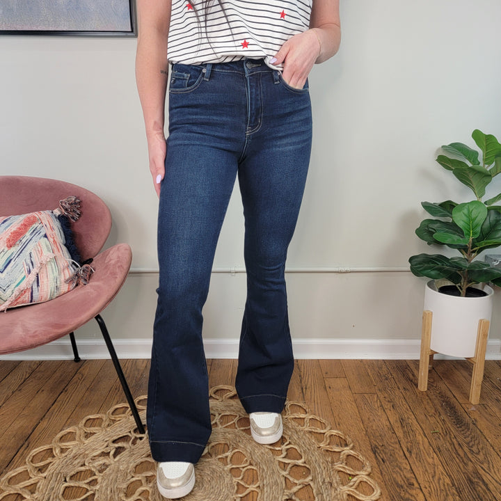 Indoors, a person wears KanCan’s Maven High Rise Flares in Dark Wash, paired with a white sleeveless top with red and blue stars and white sneakers. A pink chair and potted plant appear in the background.