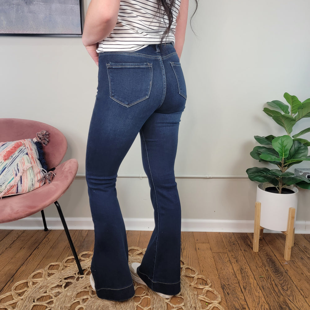 Someone wearing Maven High Rise Flares from KanCan stands on a circular rug in a room featuring a pink chair, patterned pillow, potted plant, and light-colored walls.
