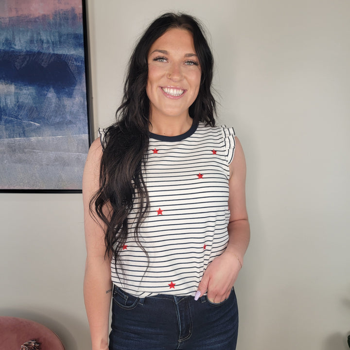 A woman with long dark hair smiles at the camera, wearing the Doe & Rae Katya Star Embroidered Top—navy and white striped with red stars—and dark jeans. Abstract artwork hangs on the wall behind her.