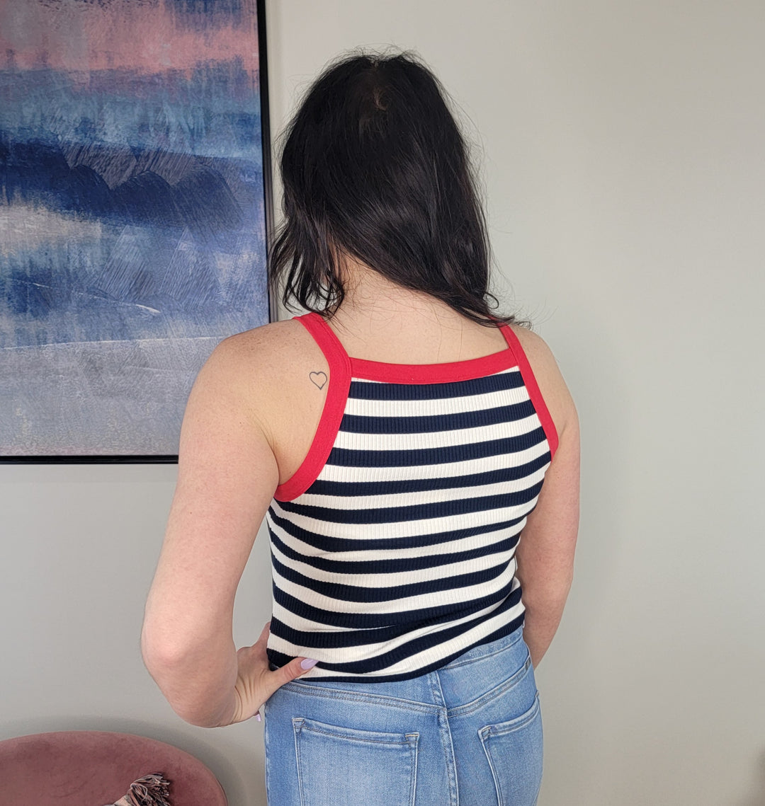 A person with long dark hair, wearing the Doe &amp; Rae Damira Striped Tank and blue jeans, stands facing away from the camera in a minimally decorated room.