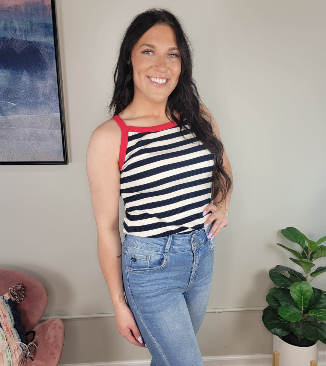 A smiling woman with long dark hair wears the Damira Striped Tank by Doe & Rae, featuring red trim, paired with high-waisted blue jeans. She stands indoors near a green plant and a framed picture on the wall.