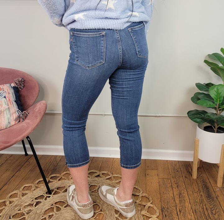 A person wearing Silvie Capris by Judy Blue and light sneakers stands on a woven rug in a room with wood floors, near a pink chair with a pillow and a potted plant. The photo, taken from behind, shows the lower half of their body.