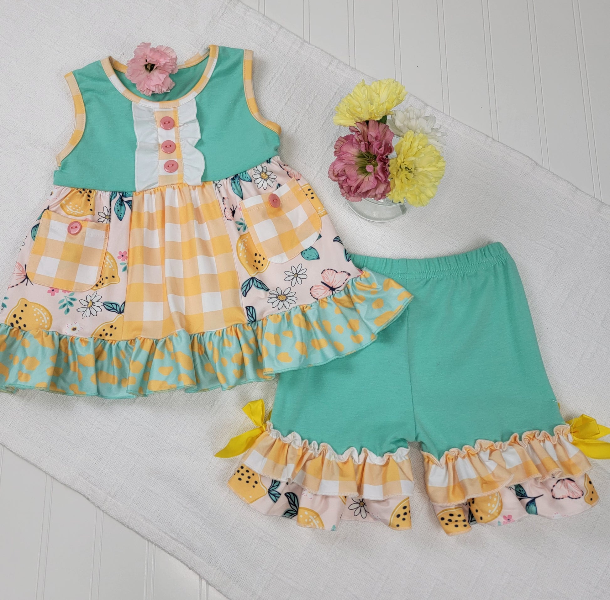 The Melina & Me Baby: Citrus Outfit is a toddler girl’s two-piece set with a sleeveless teal and yellow gingham lemon print dress with ruffles, plus matching ruffle-hem teal shorts, displayed on a white textured surface.