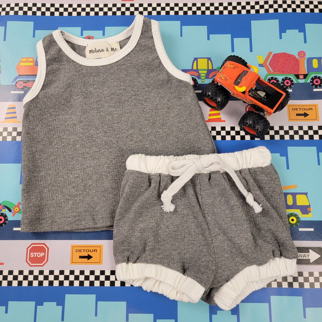 The Melina & Me Baby: Retro Ribbed Outfit—a gray sleeveless top and elastic waist shorts with white trim—is displayed on a colorful toy-themed mat next to an orange toy monster truck.