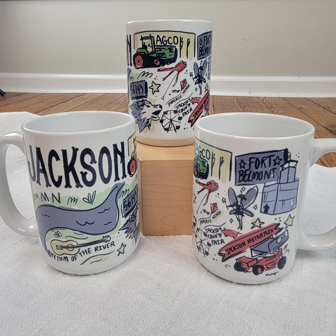 Three Ivory and Sage Jackson Mugs with colorful, hand-drawn designs about Jackson, Minnesota are shown on a white surface; one is elevated on a wooden block, while two are positioned in front and to the sides.