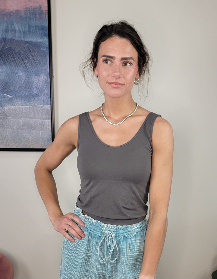 A woman with dark hair in a loose updo wears Yelete Group’s Wide Strap Layering Tank - Shorter Length in gray, blue drawstring pants, and a pearl necklace, standing indoors by a framed abstract painting.