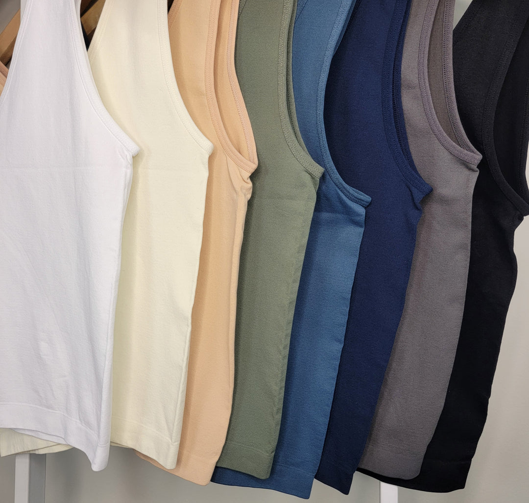 Seven Yelete Group Wide Strap Layering Tanks - Shorter Length, in white, cream, peach, olive green, teal, navy blue, gray, and black hang on wooden hangers in a row against a white background.