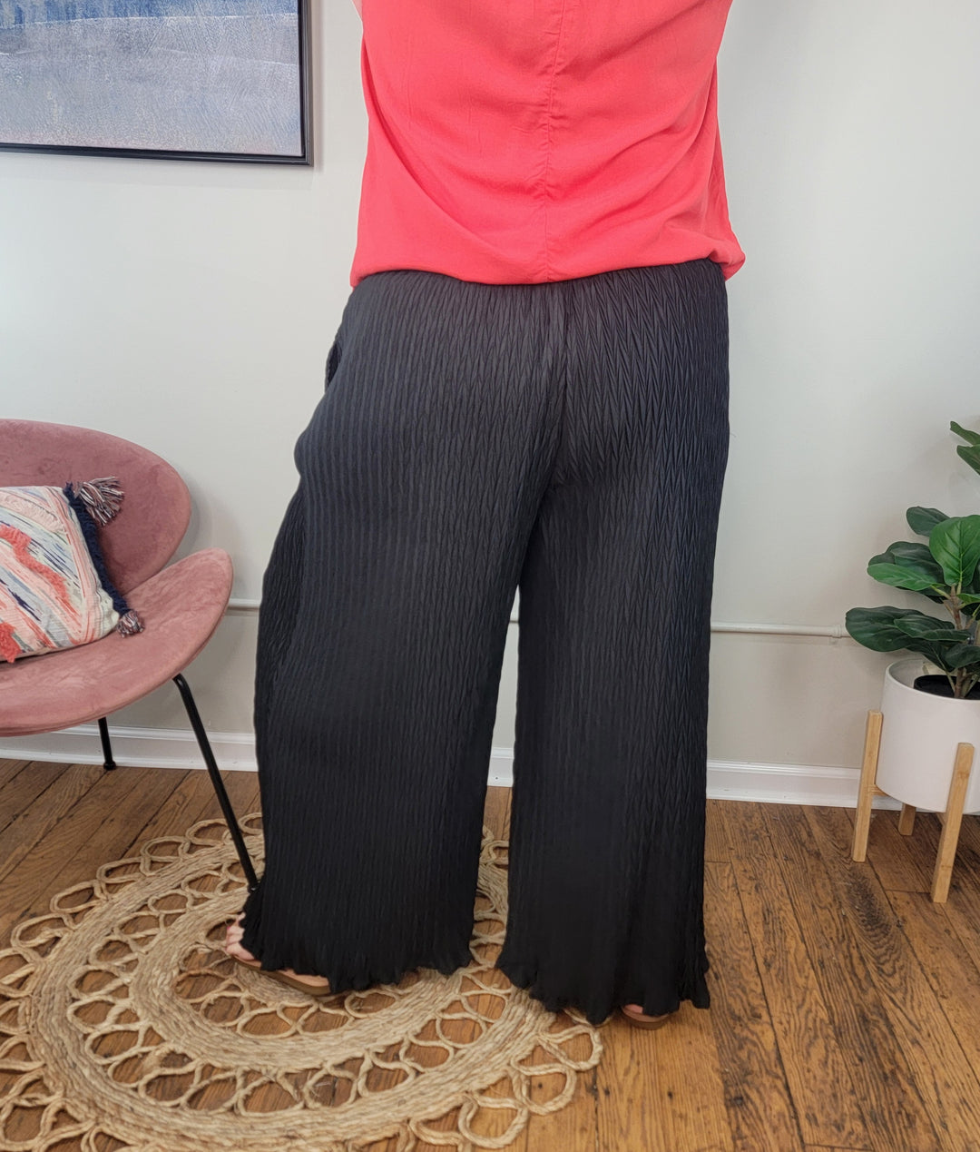 A person stands indoors on a woven rug wearing Heyson’s Karen Dress Pants with an elastic waistband and a bright coral top. The room has wooden floors, a pink chair with a pillow, framed art, and a potted plant.