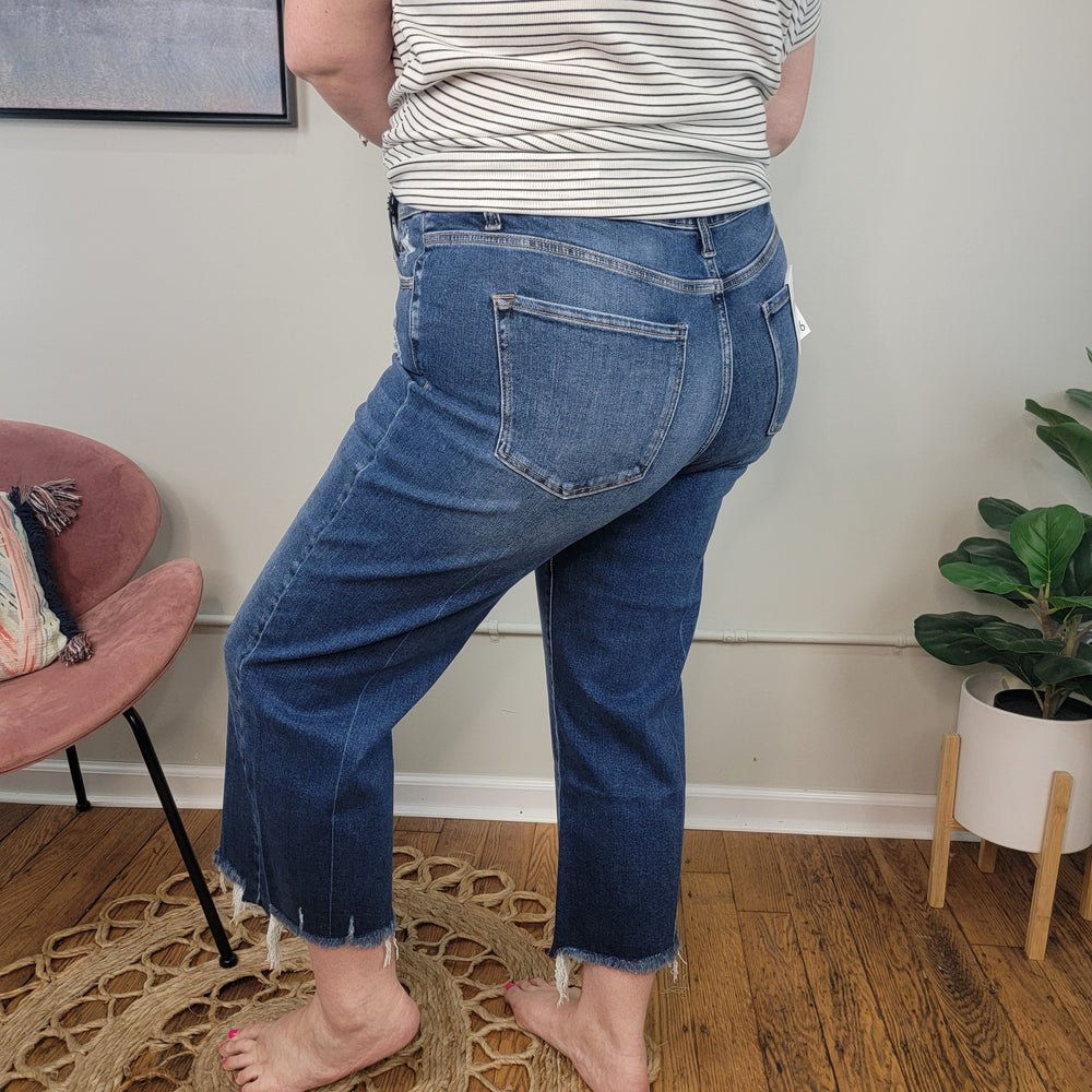 A person stands indoors on a woven rug, wearing Zenana’s Ceci Cropped Straight Jeans with a frayed hem and a striped shirt. In the background, there’s a pink chair with a pillow, a framed picture, and a potted plant on the wooden floor.