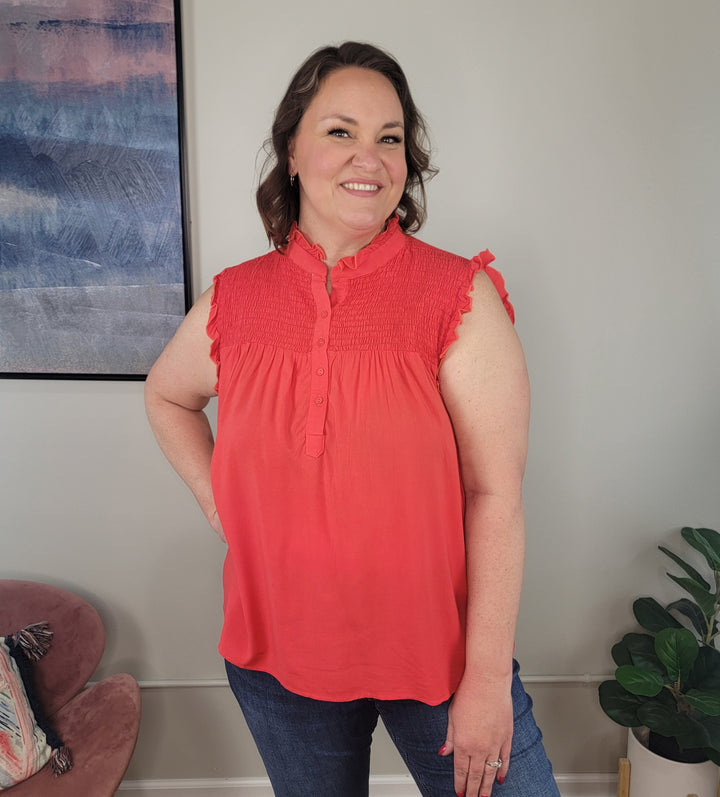 A woman with shoulder-length brown hair smiles indoors, wearing the Amaya Sleeveless Top by Culture Code and jeans. Behind her are a framed abstract painting, a pink chair, and a green plant.