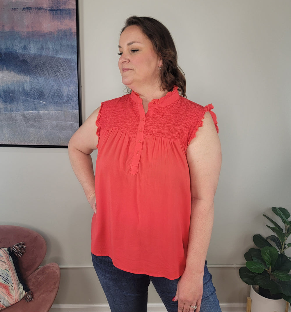 A woman models the Culture Code Amaya Sleeveless Top in coral with blue jeans, standing beside a pink cushioned chair, framed abstract art, and a potted plant.