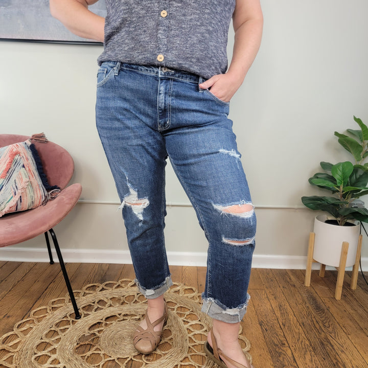 Wearing a gray sleeveless top and KanCan's Tara Slim Boyfriend Jeans, a person stands on a woven rug with one hand in their pocket. Behind them, a pink chair with a striped pillow and a green plant rest on the wooden floor.