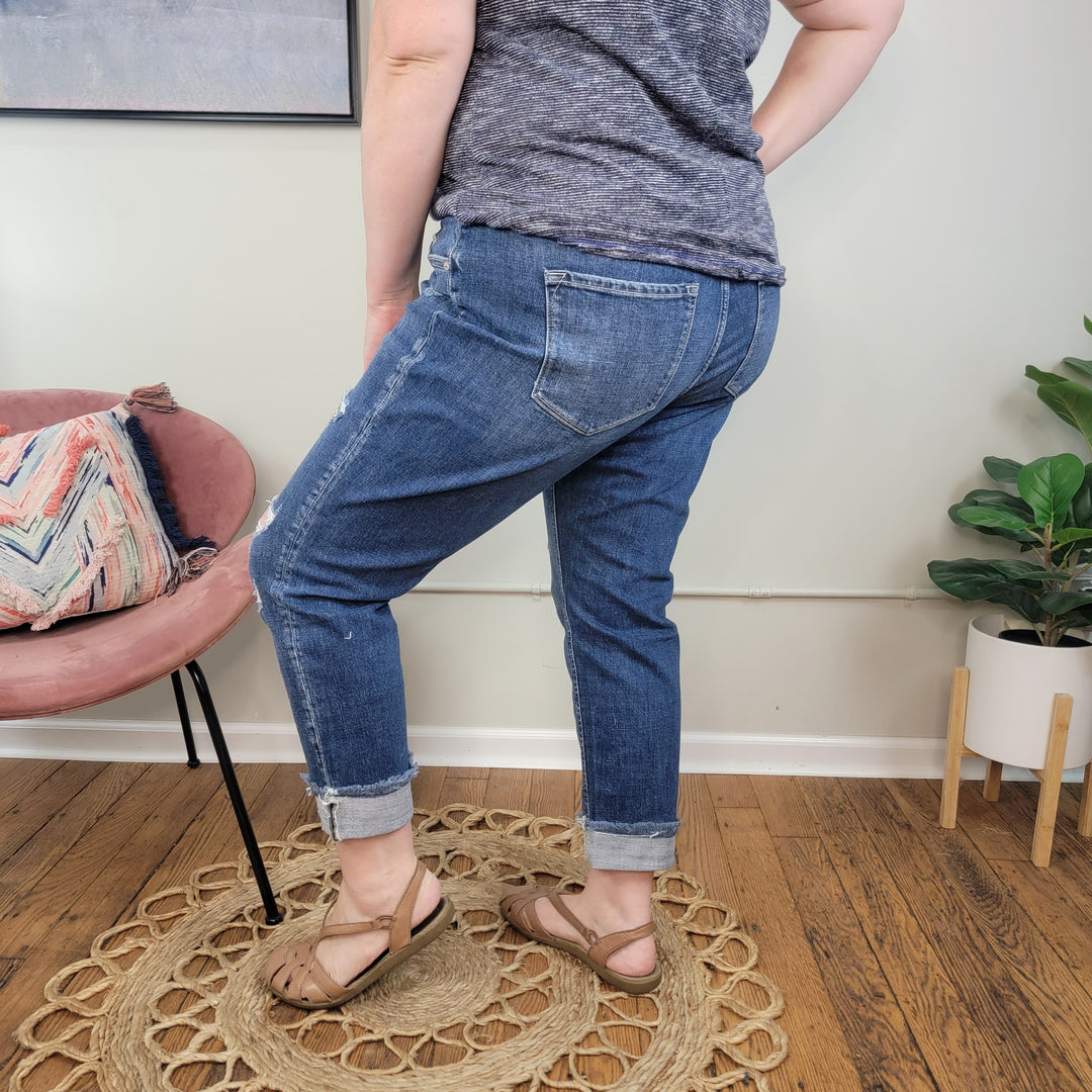 Indoors, a person wears Tara Slim Boyfriend Jeans from KanCan, a gray shirt, and sandals while standing on a circular woven rug. Nearby are part of a pink chair with a colorful pillow and a potted plant.