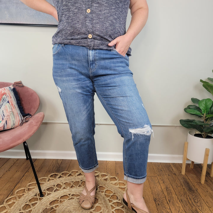 A person stands indoors on a braided rug wearing a gray short-sleeve top and KanCan's Stacie Boyfriend Jeans, featuring patched distressing. They have tan sandals, hand in pocket, with a pink chair and leafy plant nearby.