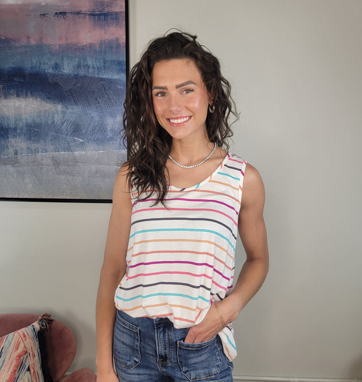 A woman with wavy brown hair stands indoors, smiling at the camera. She wears the Haptics Meara Stripe Tank and blue jeans, with a large abstract painting visible on the wall behind her.