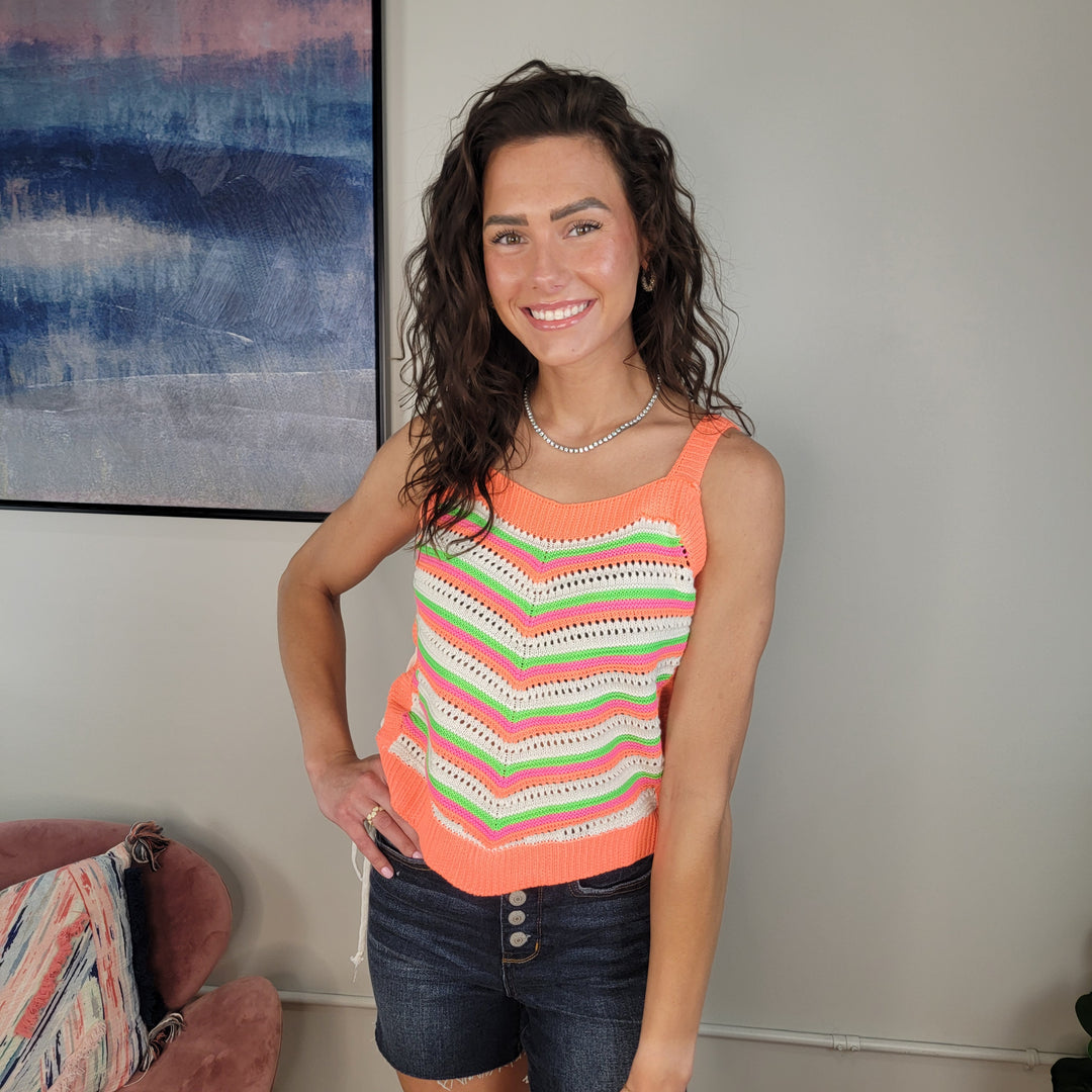A woman with curly dark hair smiles at the camera, wearing the Kimia Crochet Tank by Davi & Dani, featuring colorful stripes and lace-up sides, paired with dark denim shorts. She stands indoors near a pink chair and an abstract painting.
