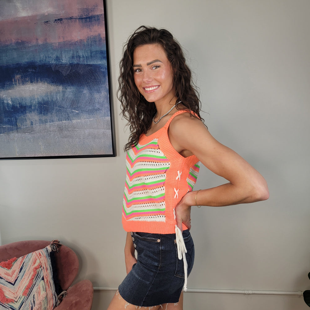 A smiling woman with curly hair models the Kimia Crochet Tank by Davi & Dani, featuring lace-up sides, paired with a dark denim skirt. She stands sideways in a modern room with abstract art and a pink chair in the background.