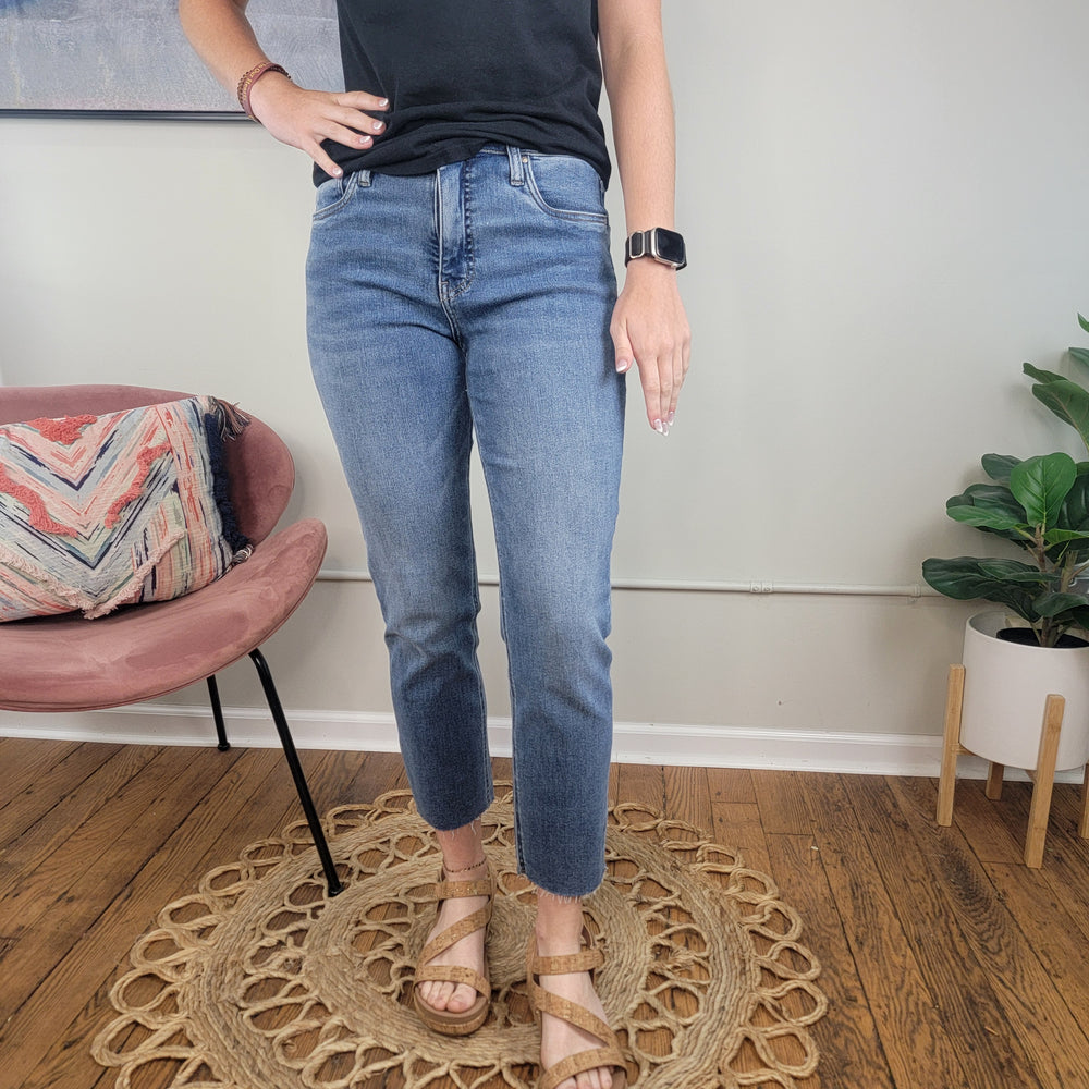 Someone stands indoors on a round woven rug wearing Rachael Mom Jeans from Kut by Kut from the Kloth, a black top, a smart watch, and tan sandals. A pink chair with a patterned pillow and green plant are in the background.