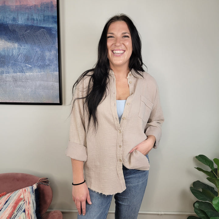 A woman with long dark hair smiles indoors, wearing the Zenana Aimee Gauze Button Down over a white top and blue jeans, standing beside a pink chair, colorful pillow, leafy plant, and abstract wall art.