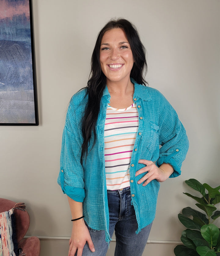 A woman with long dark hair smiles indoors, wearing the Zenana Aimee Gauze Button Down in turquoise over a striped top and jeans. A framed picture decorates the wall, and a potted plant sits beside her.