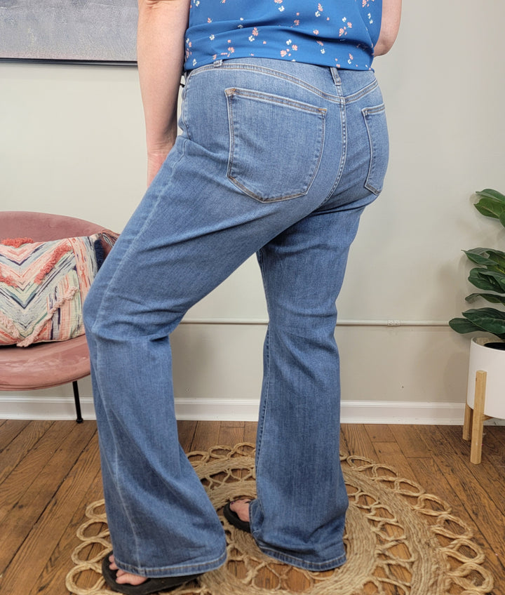 A person wearing the Darcie Flares from Judy Blue and a blue floral top stands on a circular woven rug in a room with wooden floors, a pink chair, a plant, and a beige wall. They are wearing black sandals.