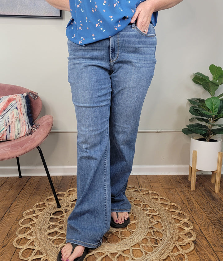A person wearing light blue Darcie Flares from Judy Blue, a blue floral top, and black sandals stands on a woven rug. A pink chair with a patterned pillow and a green potted plant are visible in the background.
