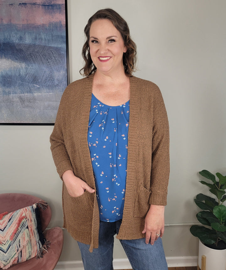 A woman with wavy brown hair smiles in a living room, wearing the ee:some Honor Cardigan over a blue floral blouse. Modern decor includes a stylish chair, cushion, abstract wall art, and a green plant.