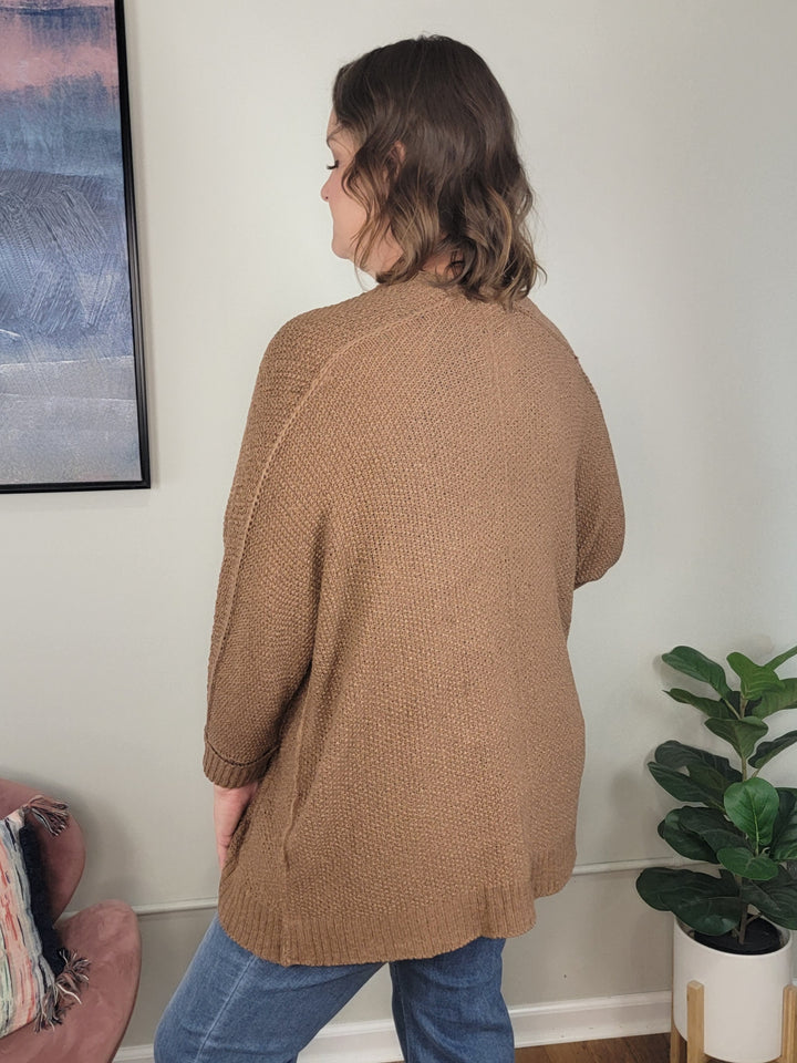 A woman with wavy brown hair stands indoors with her back to the camera, wearing the ee:some Honor Cardigan and blue jeans. A plant and framed artwork nearby complete this perfect inspiration for fall layering sweater looks.
