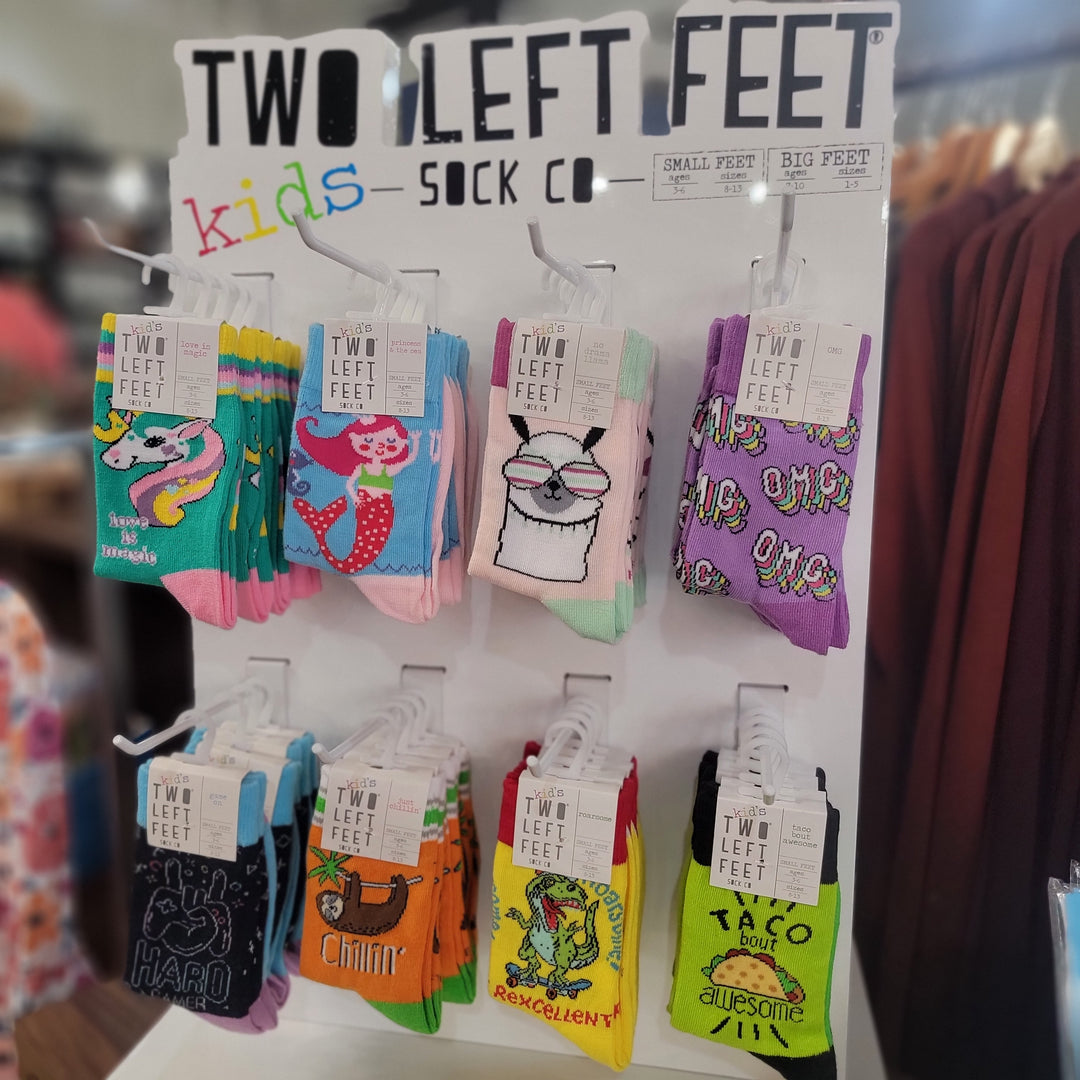 A display rack of Two Left Feet Socks by DM Merchandising, featuring printed socks for kids with designs like unicorns, mermaids, tacos, and cartoon characters. Each pair is hung on hooks with labeled tags and kids sock sizes.