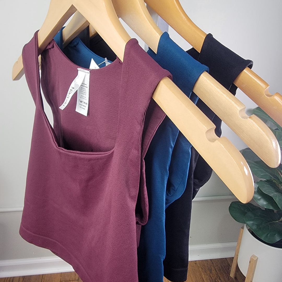 Four NikiBiki Square Neck Crop Tops in burgundy, navy blue, teal, and black by NikiBiki hang on wooden hangers, with a small white-potted plant in the background.
