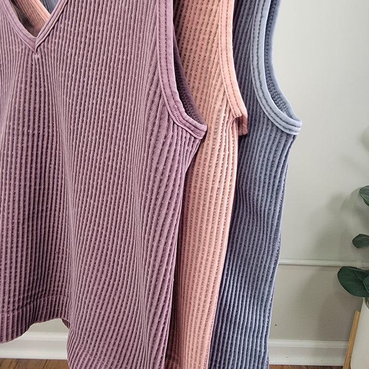 Three NikiBiki Reversible Ribbed Crop Tops in mauve, blush pink, and gray hang on wooden hangers against a light wall with a green plant. Each one-size-fits-most top features a reversible neckline. Brand: NikiBiki.