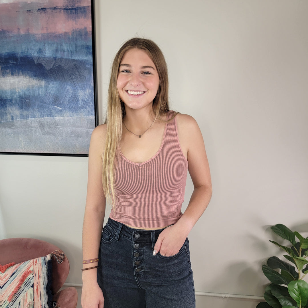 A young woman with long blonde hair smiles indoors near a plant and wall art, wearing the NikiBiki Reversible Ribbed Crop Top in pink by NikiBiki with high-waisted dark jeans.