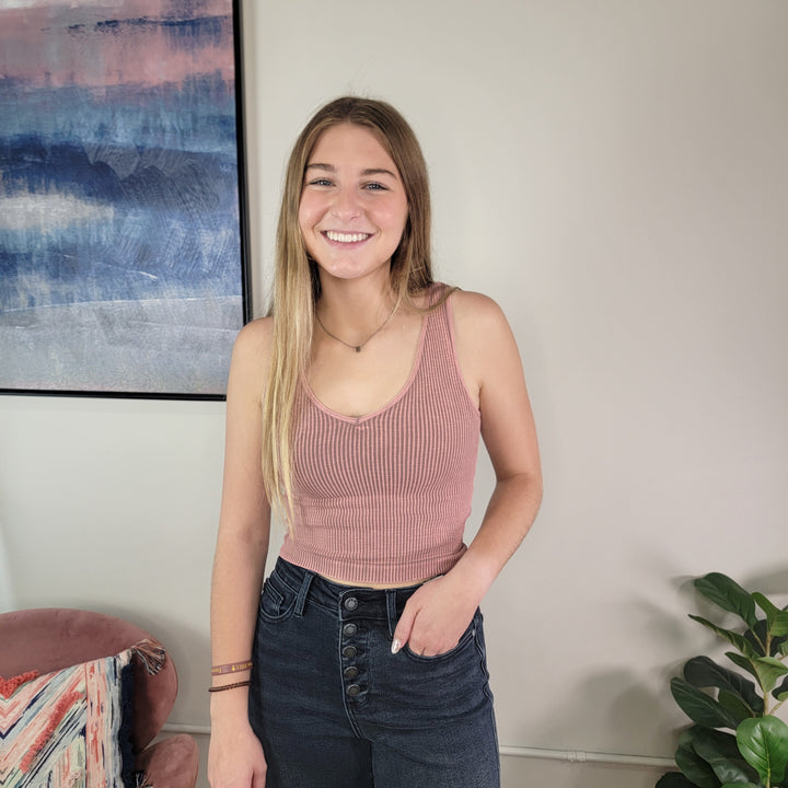 A young woman with long blonde hair smiles indoors near a plant and wall art, wearing the NikiBiki Reversible Ribbed Crop Top in pink by NikiBiki with high-waisted dark jeans.