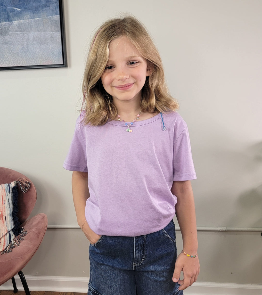 A young girl with shoulder-length blonde hair smiles at the camera, wearing the Cutie Patootie Girls: Ella Tee in light purple, blue jeans, a beaded necklace, and bracelet. She stands indoors with one hand in her pocket.