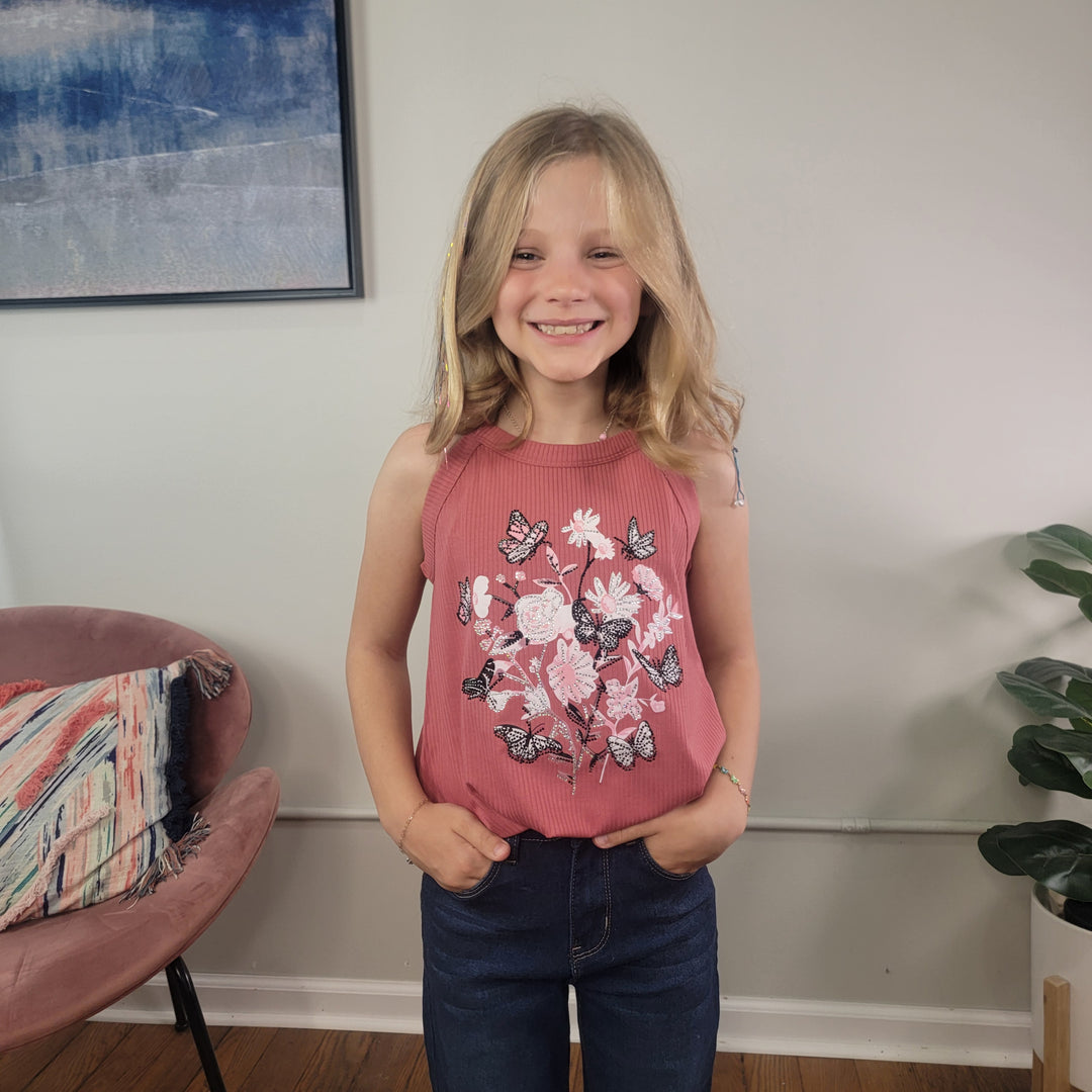 A smiling young blonde girl stands indoors wearing the Cutie Patootie Girls: Thea Tank—a mauve ribbed tank top with a white floral and glitter butterfly print—paired with dark jeans. Behind her are a pink chair, patterned pillow, wall art, and a green plant.