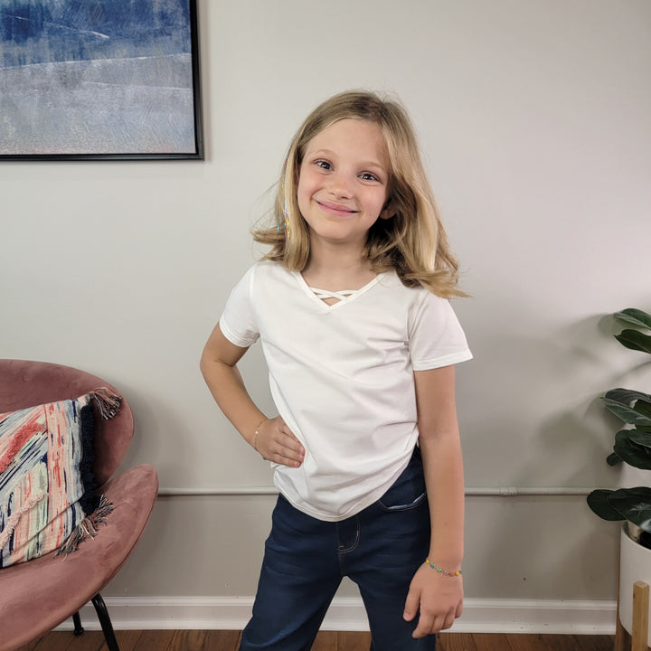 A smiling young girl with blonde hair stands indoors, wearing the Cutie Patootie Girls: Karli Tee in white and blue jeans. She poses with a hand on her hip. Behind her are a pink chair, colorful cushion, plant, and framed wall art.