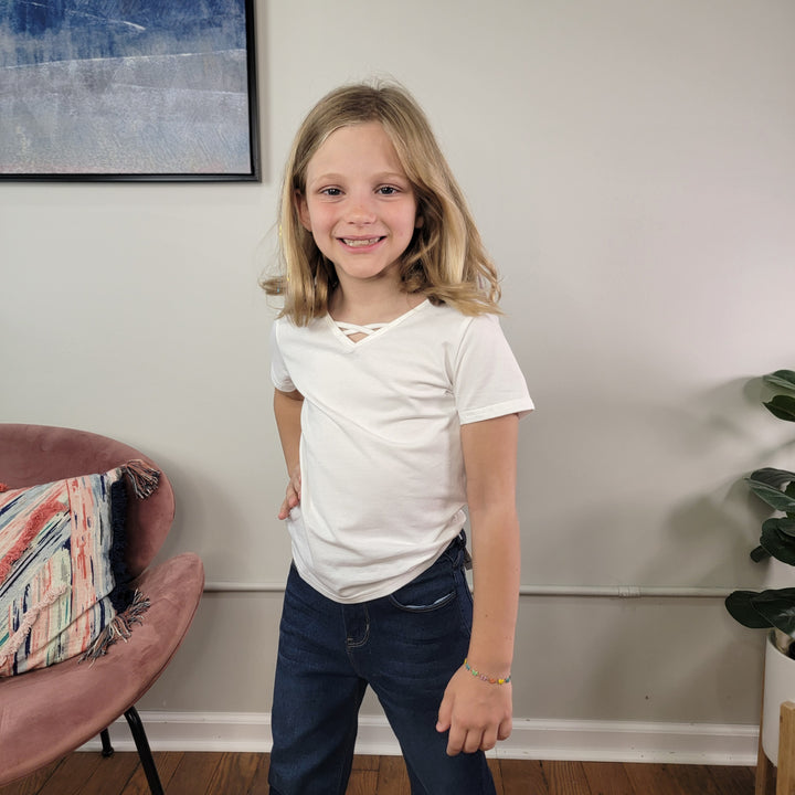 A young blonde girl smiles at the camera, wearing the Cutie Patootie Girls: Karli Tee with blue jeans. She stands indoors by a pink chair, a plant, and a framed wall picture.