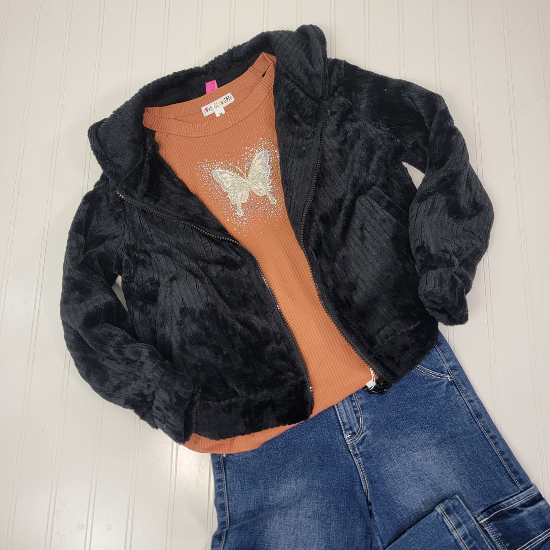 A flat lay of a child's outfit with a black faux fur jacket, Cutie Patootie's Beatrice Tank in cinnamon with a glitter butterfly patch, and blue denim jeans, all arranged on a white textured surface.