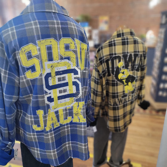 Two mannequins sport Game Day Social’s SDSU Plaid shirts—one in blue with yellow and white SDSU Jacks text, the other in yellow and black with SIDWAK and a mascot—perfect for showing school spirit indoors.