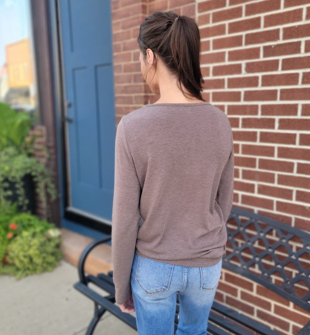 A woman with brown hair in a ponytail, wearing the Staccato Denise Top in mocha and blue jeans, stands outside facing away from the camera near a brick wall, a blue door, and a black metal bench.