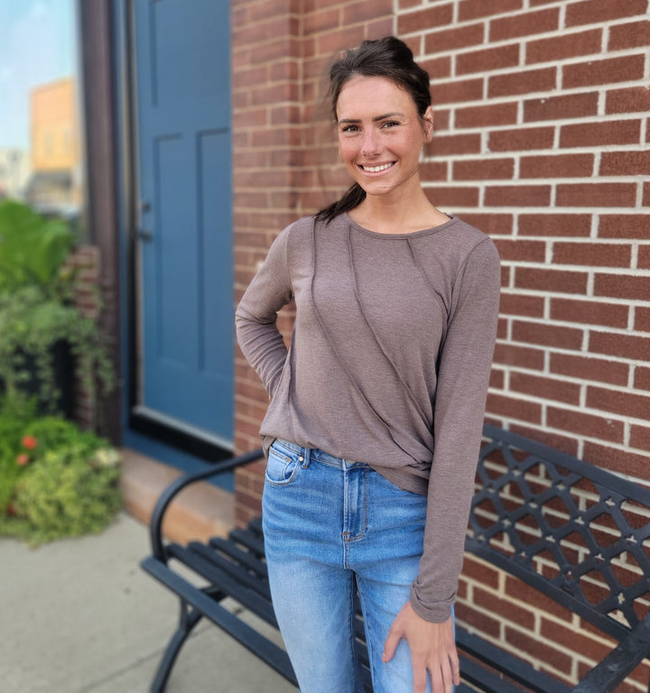 A woman with long brown hair smiles beside a black metal bench, wearing the Staccato Denise Top in mocha and blue jeans, standing in front of a brick wall and blue door with greenery in the background.