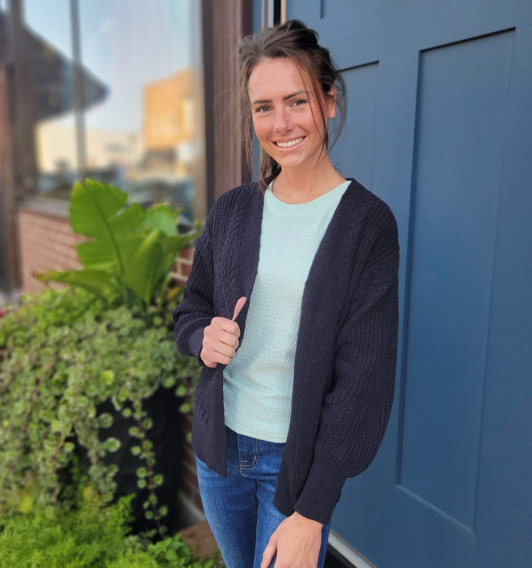A woman with brown hair, smiling in front of a blue door and green plants, wears the Annva Fashion Virginia Cable Knit Cardigan over a light blue top.