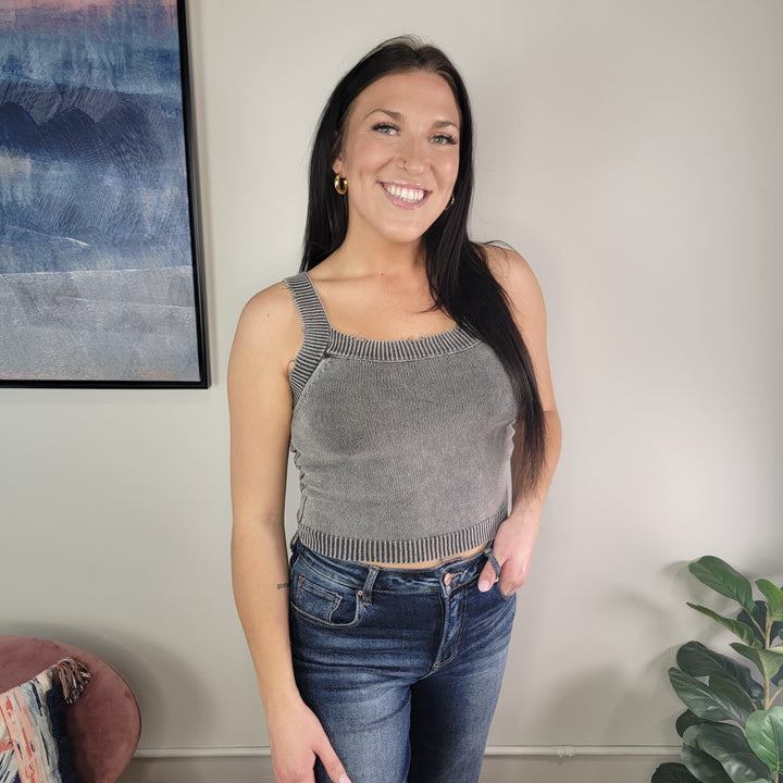 A woman with long dark hair wears the Tiffany Knit Tank Top by Timing and blue jeans, smiling in a well-lit room with a large abstract painting, pink chair, and green plant in the background.