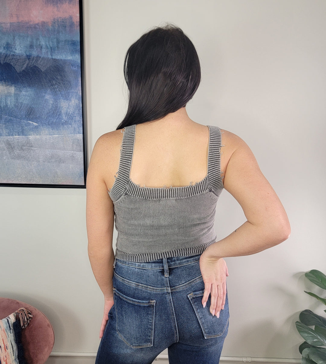 A woman with long dark hair stands facing away, wearing the charcoal Tiffany Knit Tank Top by Timing with blue jeans. One hand is on her hip. A framed abstract artwork and a plant appear in the background.