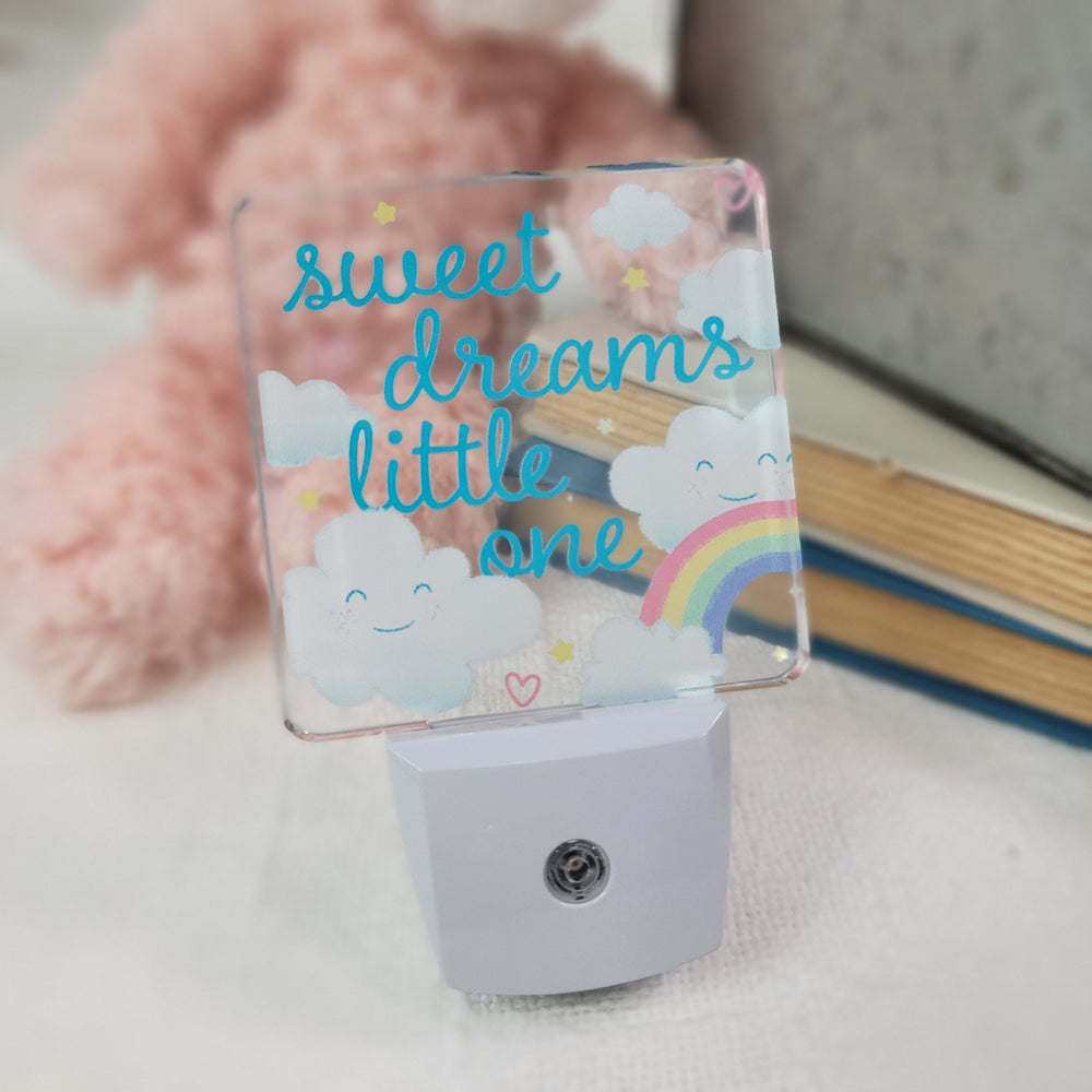 Fosters Nightlights feature smiling clouds, a rainbow, and stars with “sweet dreams little one” in blue and yellow. Part of the Nightlights collection, it’s shown beside a soft pink plush toy and blurred books in the background.