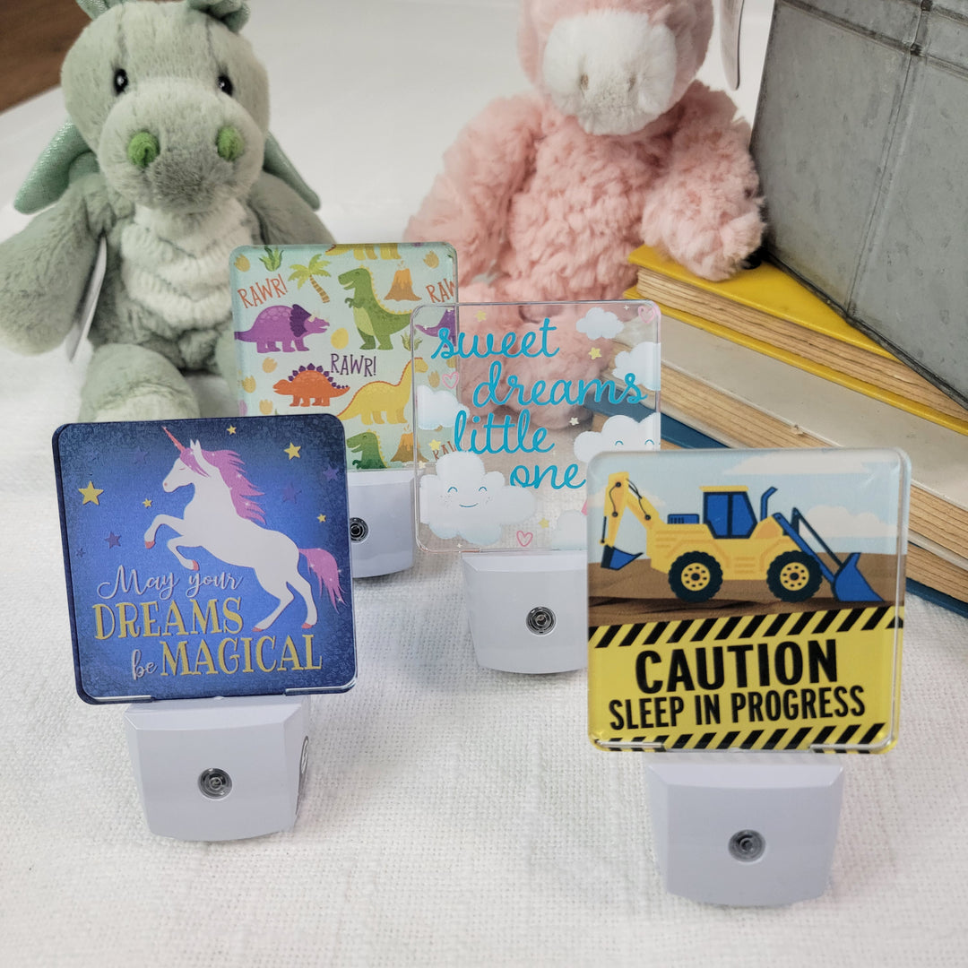 Four Fosters Nightlights with unique designs and messages are displayed on a white surface, with two plush toys—a green dragon and a pink dinosaur—and books in the background.