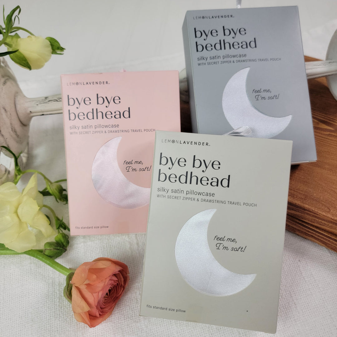 Three DM Merchandising Satin Pillowcases in pink, gray, and silver are shown on white fabric with flowers and a tray—ideal for taming frizzy hair. Each box reads, "feel me, I'm soft!.