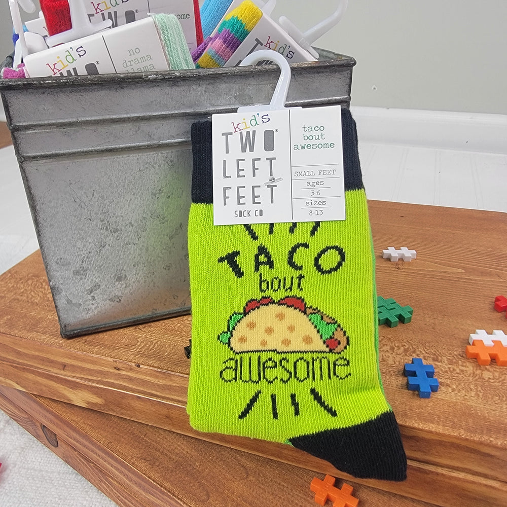 A pair of Two Left Feet Socks by DM Merchandising for kids, in green and black with a taco illustration and "Taco bout awesome" text, hang from a fun socks box in the background, with puzzle pieces scattered nearby.